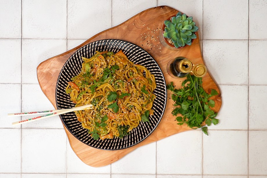 Black and white bowl on a wooden board piled with vegetable satay noodles with chopsticks and herbs