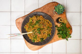 Black and white bowl on a wooden board piled with vegetable satay noodles with chopsticks and herbs