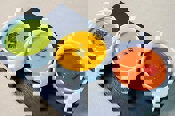 Three small white dishes of soup, one green with a broccoli topping, one orange with a topping of pumpkin seeds and one red with a topping of tomato. Photo by Farhad Ibrahimzade